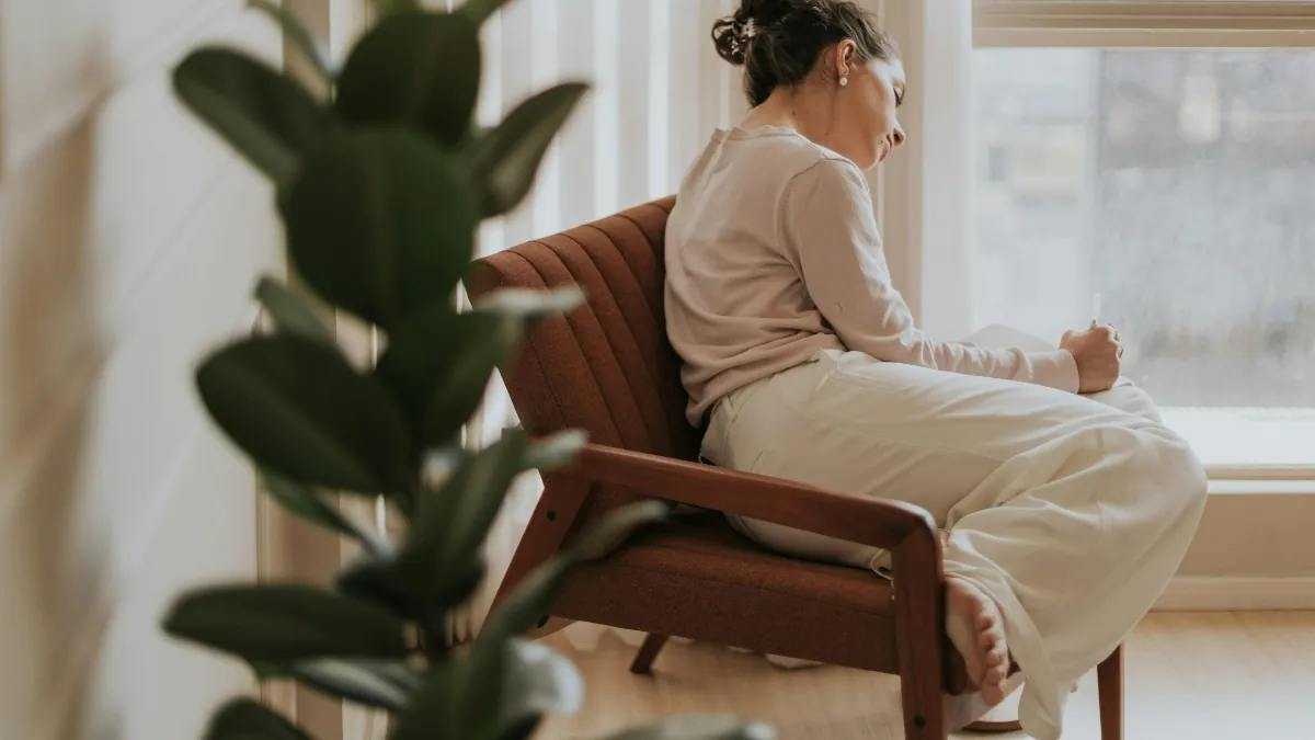 Mulher madura ao lado da janela com expressão serena.